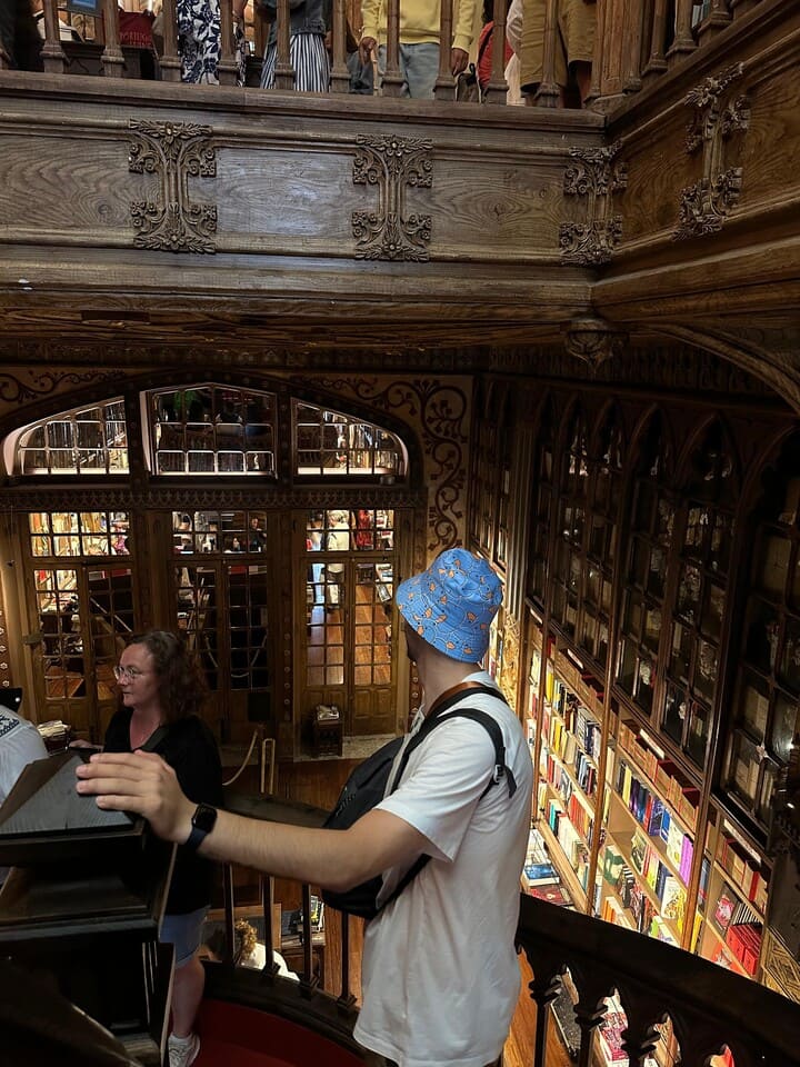 Uomo con cappello azzurro a fantasia e zaino nero mentre osserva i dettagli in legno della celebre libreria Livraria Lello di Porto, affacciato dalla scala interna.