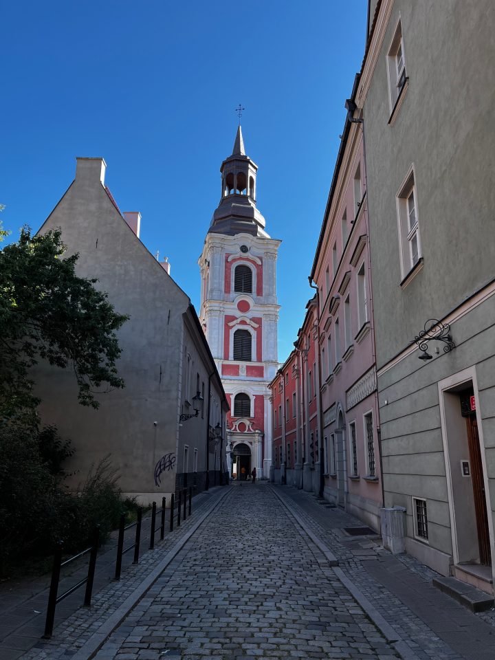 La Fara Poznańska, Chiesa di San Stanislao a Poznań, vista da una stradina acciottolata che conduce alla sua facciata barocca rosa e bianca.