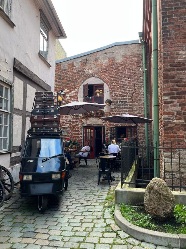 Locale caratteristico a Riga con cortile in mattoni rossi, tavolini all’aperto e Ape Piaggio decorata con valigie, ideale per una pausa caffè nel centro storico.