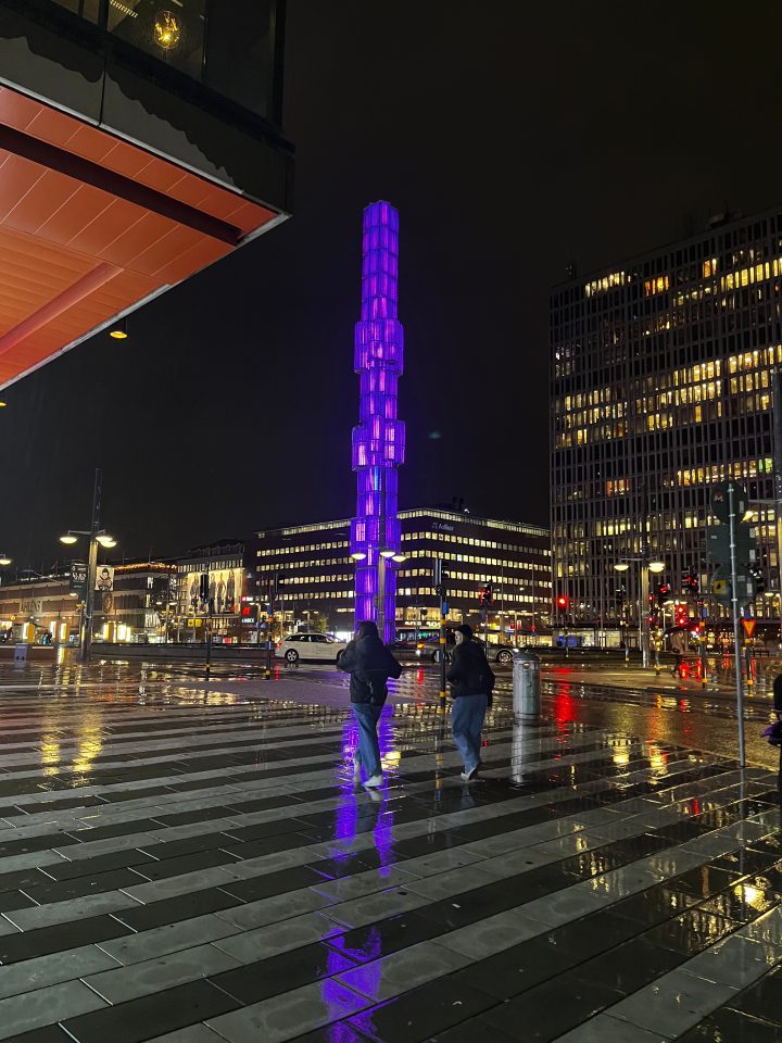 Sergels Torg a Stoccolma di notte, con la torre di vetro illuminata di viola e i riflessi sul pavimento bagnato.